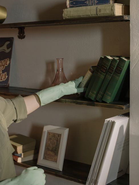 Close-up of a person's arm wearing a light green cleaning glove, reaching towards a wooden bookshelf with a dark finish, holding several neatly stacked white paper documents or magazines. The bookshel