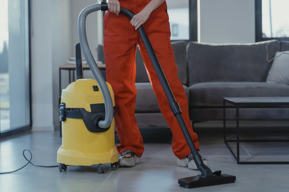 A person dressed in orange overalls is operating a yellow commercial carpet cleaner with a black hose attachment in a modern living room. The room features a large window letting in natural light, a dark gray sofa with light-colored cushions, and a black metal coffee table on a clean, tiled floor. The cleaning process appears thorough, with the vacuum head in contact with the floor surface, reflecting surface cleaning and deep cleaning undertaken by Carpet Cleaners SE11 in accordance with their residential and commercial cleaning services, focusing on hygiene and maintenance in the Kennington Park area.