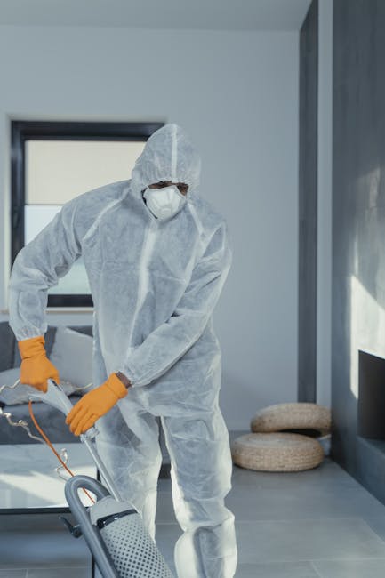 A professional cleaner dressed in full protective gear, including a white disposable coverall, face mask, safety goggles, and orange gloves, operating a vacuum cleaner on a light-colored tiled floor in a modern, well-lit room with gray walls and large windows. The room appears clean and tidy, with natural light streaming in, illuminating the polished surfaces and creating a bright atmosphere. The image emphasizes surface cleaning, deep cleaning, and sanitisation processes as provided by Carpet Cleaners SE11 in the Kennington Park area. The setup suggests a focus on thorough domestic cleaning, ensuring hygiene and maintenance of residential or commercial spaces.