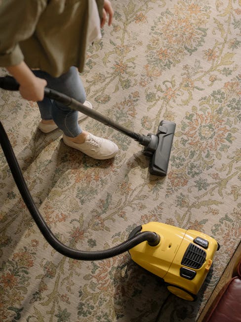 A person dressed in orange overalls is operating a yellow commercial carpet cleaner with a black hose attachment in a modern living room. The room features a large window letting in natural light, a dark gray sofa with light-colored cushions, and a black metal coffee table on a clean, tiled floor. The cleaning process appears thorough, with the vacuum head in contact with the floor surface, reflecting surface cleaning and deep cleaning undertaken by Carpet Cleaners SE11 in accordance with their residential and commercial cleaning services, focusing on hygiene and maintenance in the Kennington Park area.