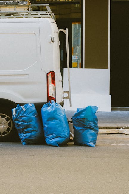 A green wheeled refuse bin labeled with the number 53 and the words 'JUN BESAR' is positioned on a concrete pavement near a street. To the left, there is a metal shelving unit with cleaning supplies a