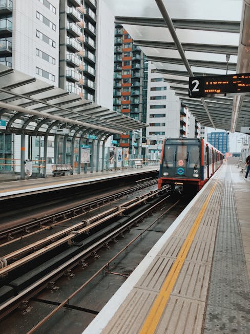 An outdoor train station platform with a modern urban backdrop of high-rise residential buildings. The platform features a tactile yellow safety line along its edge, a glass and metal shelter with ben
