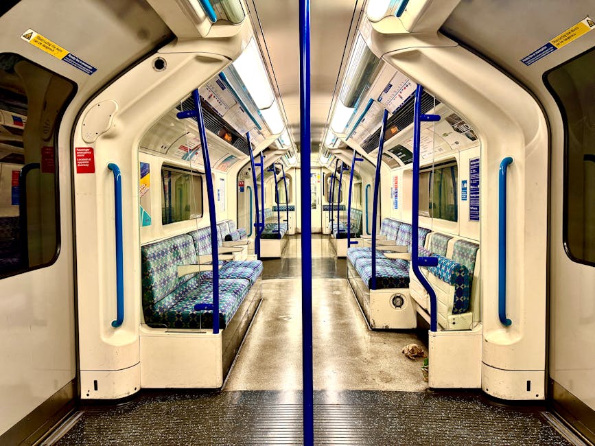 An outdoor train station platform with a modern urban backdrop of high-rise residential buildings. The platform features a tactile yellow safety line along its edge, a glass and metal shelter with ben